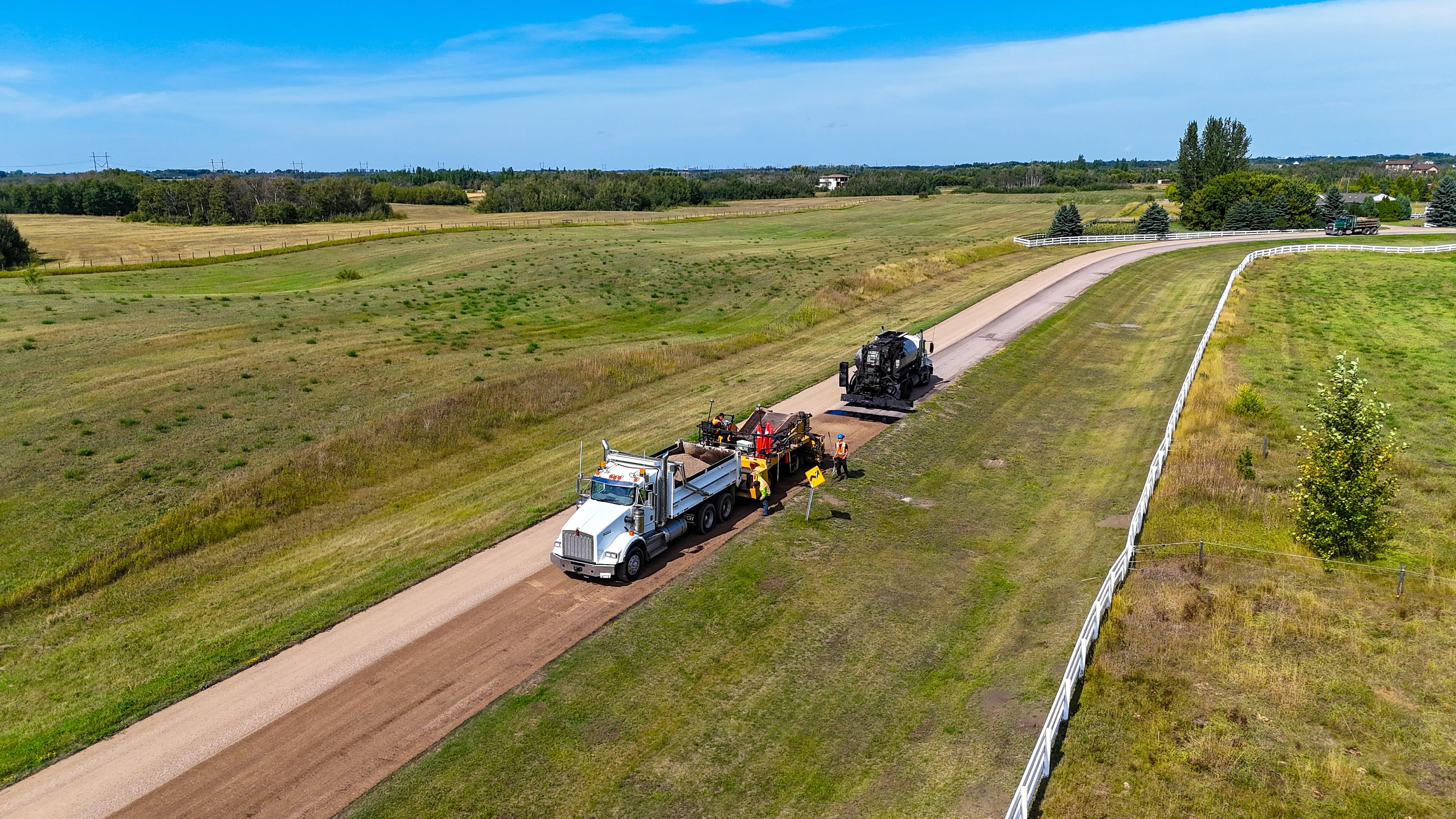 Gravel road treatment convoy on a rural municipality road with fields on both sides, seen from above